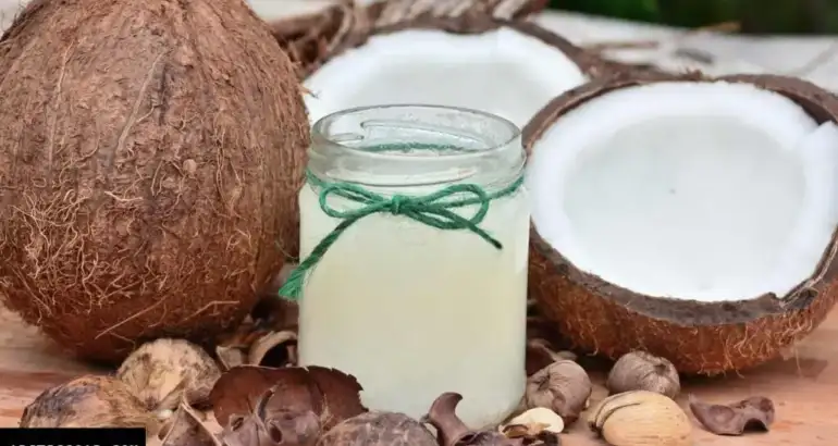 A fresh, brown hairy coconut split open on a rustic table, revealing its pure, white meat, with a jar of clear coconut oil nearby.